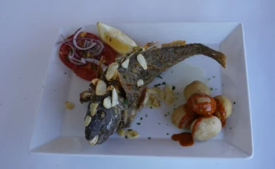Plato de pescado frito con almendras, acompañado de rodajas de tomate, cebolla morada y limón, servido con papas pequeñas y salsa.