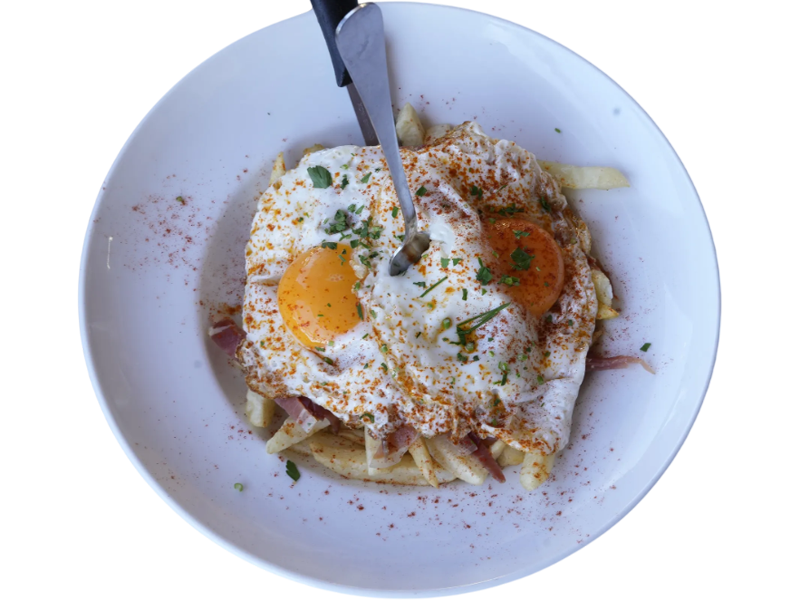 Plato de papas fritas cubiertas con jamón, dos huevos fritos y espolvoreado con pimentón y perejil fresco. Ideal para un desayuno abundante y delicioso.